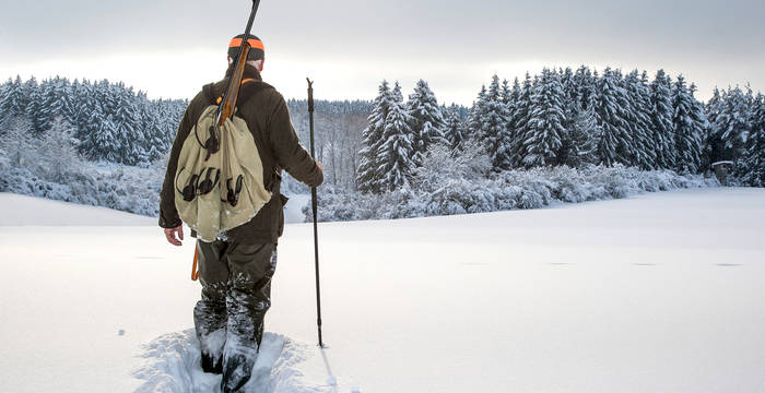 In einer Winterlandschaft geht ein Jäger durch den frischen Schnee hinunter ins Tal.