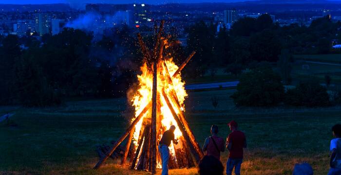 Ein brennendes 1. August-Feuer auf einem Feld mit einer Stadt im Hintergrund. Im Vordergrund sieht man ein paar Menschen.
