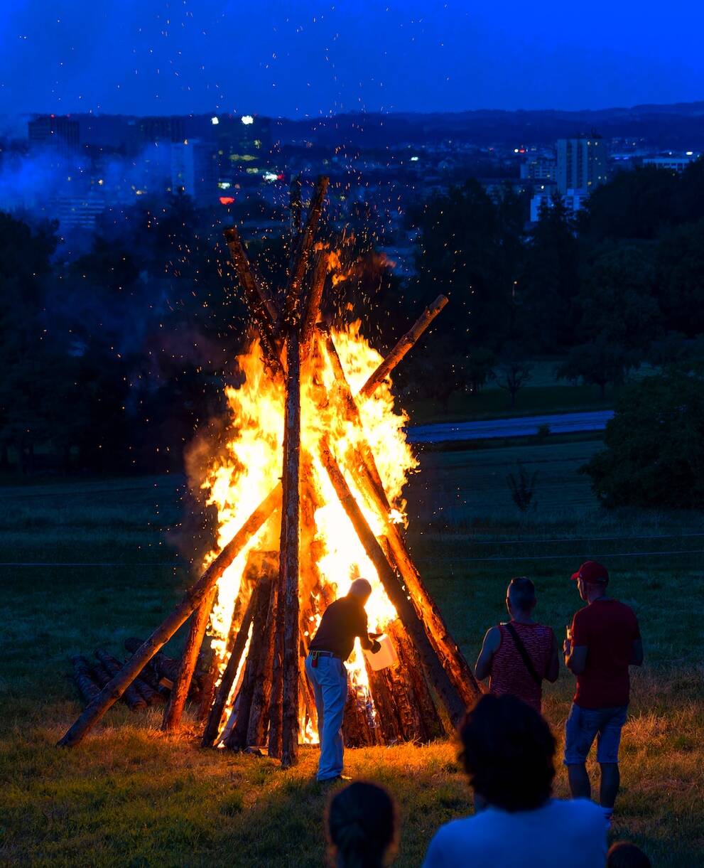 Ein brennendes 1. August-Feuer auf einem Feld mit einer Stadt im Hintergrund. Im Vordergrund sieht man ein paar Menschen.